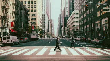 A pedestrian crossing a wide city street at a marked crosswalk, with traffic signals overhead